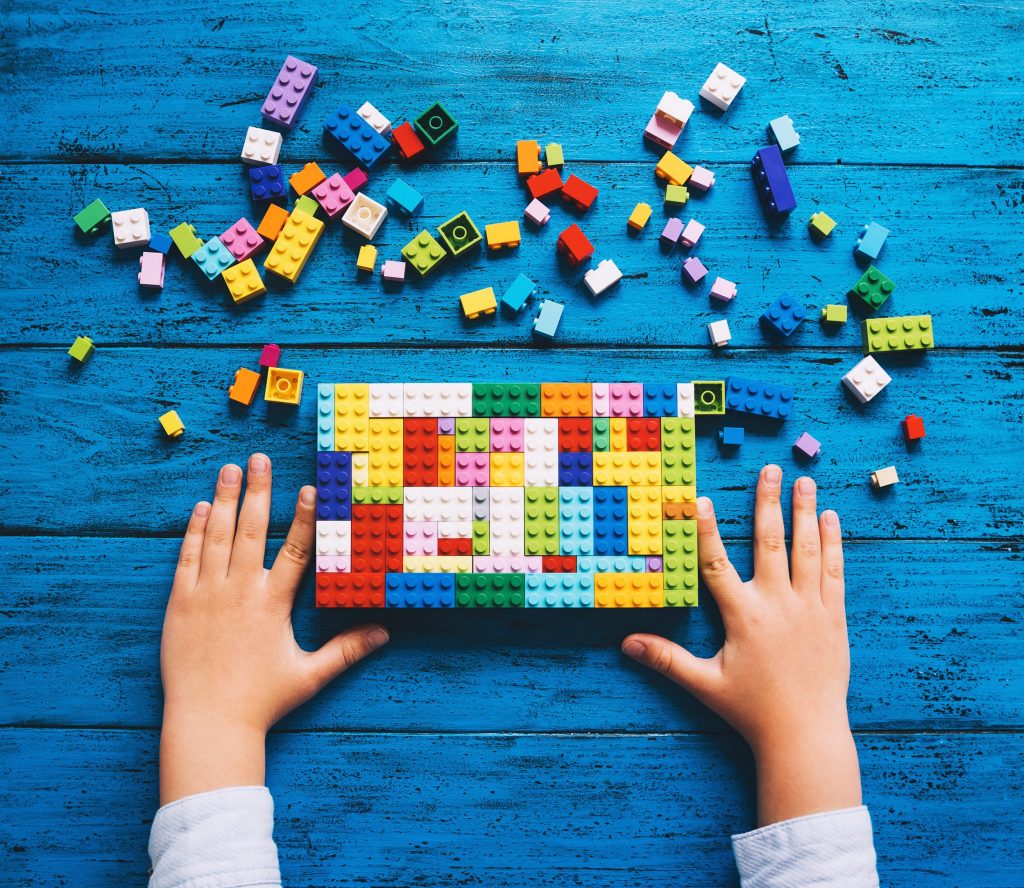 Ein paar Kinderhände, die aus vielen herumliegenden Legosteinen einen bunten Lego-Block gebaut haben.