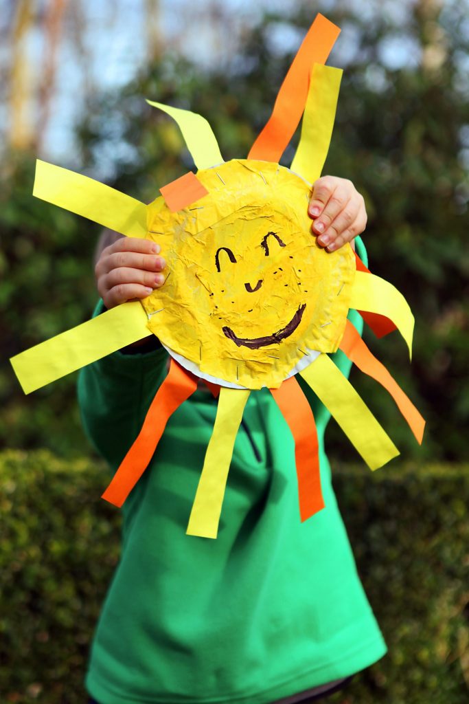 Ein kleines Kind hält eine aus Papier gebastelte, fröhlich grinsende Sonne vor sein Gesicht.